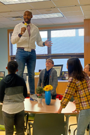 A man stands on a table, addressing a group of young adults.