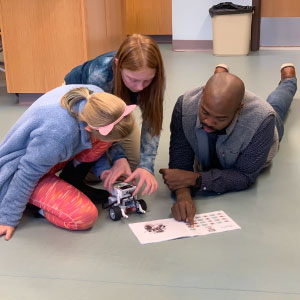 A teacher and two students sit on the floor, examining a notebook with notes together.